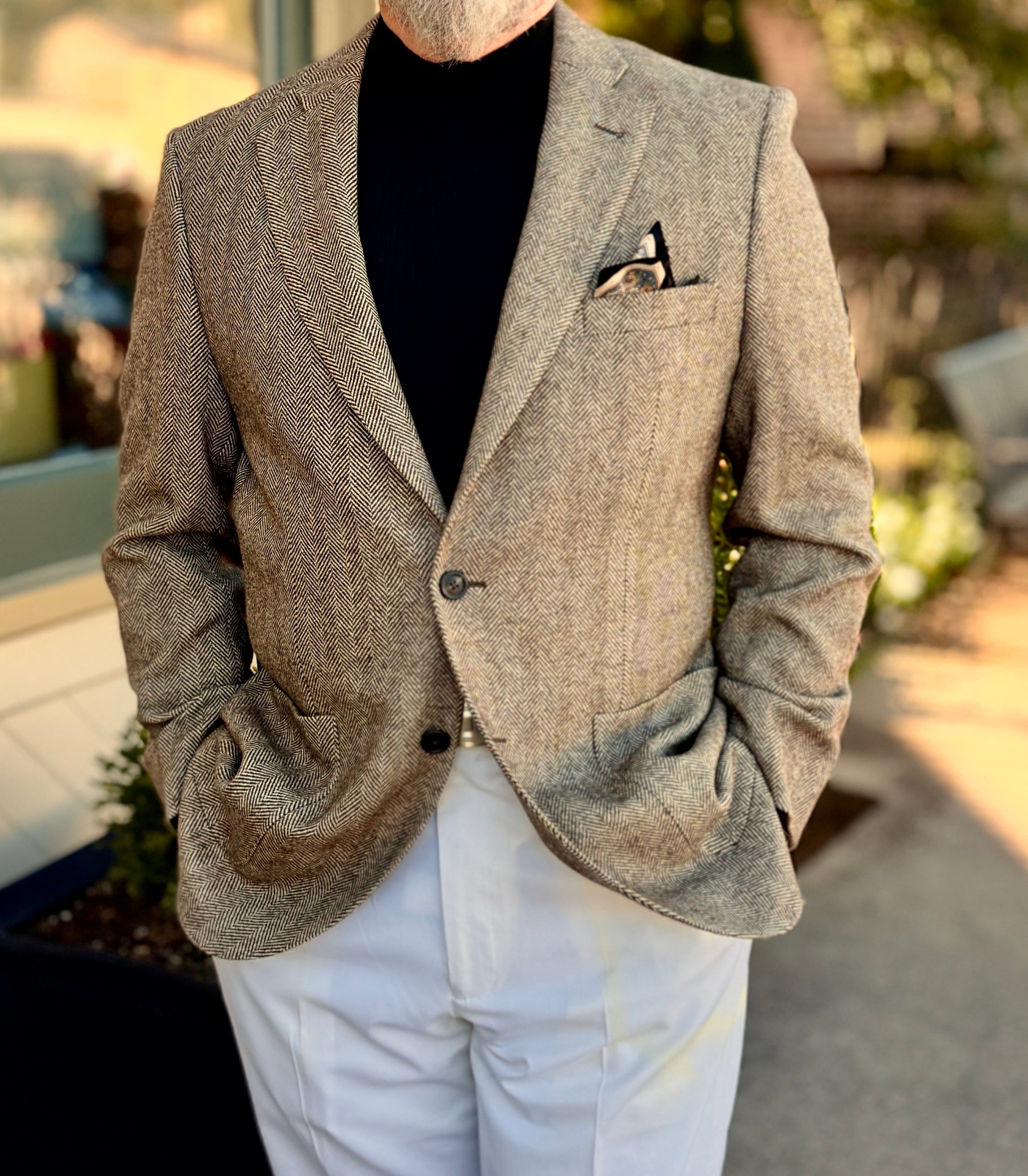 Man wearing a brown blazer over a black shirt with white pants outdoors.