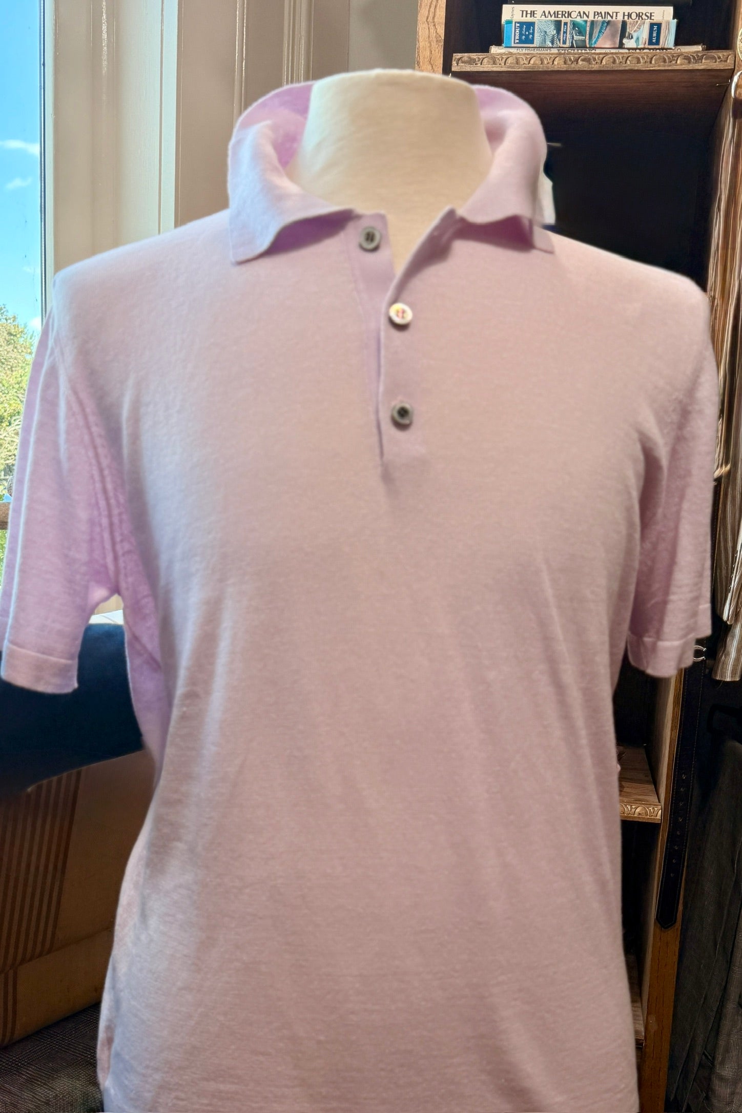 A light pink short-sleeve polo shirt displayed on a cream-colored mannequin torso, set against an indoor background with a window and wooden shelving.